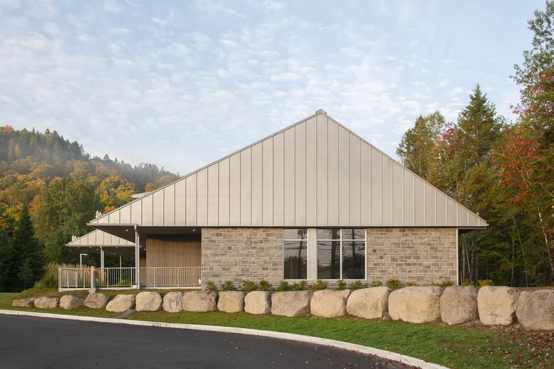 Street facade with large boulder retaining wall, tall gable, and hillside forest beyond