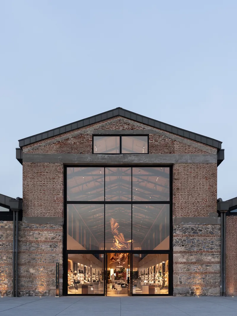 Gabled brick elevation with large glazed opening revealing timber trusses and sculptural installation inside at dusk