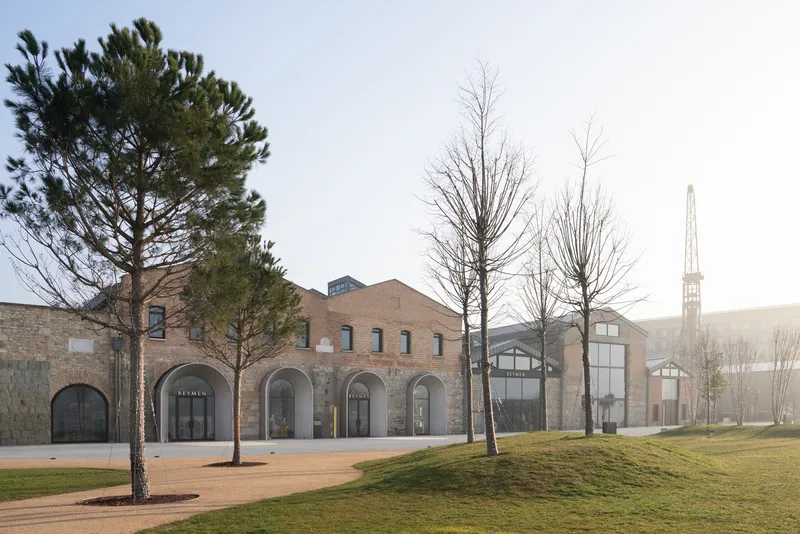 Brick facade with arched openings facing a gravel plaza lined with bare winter trees