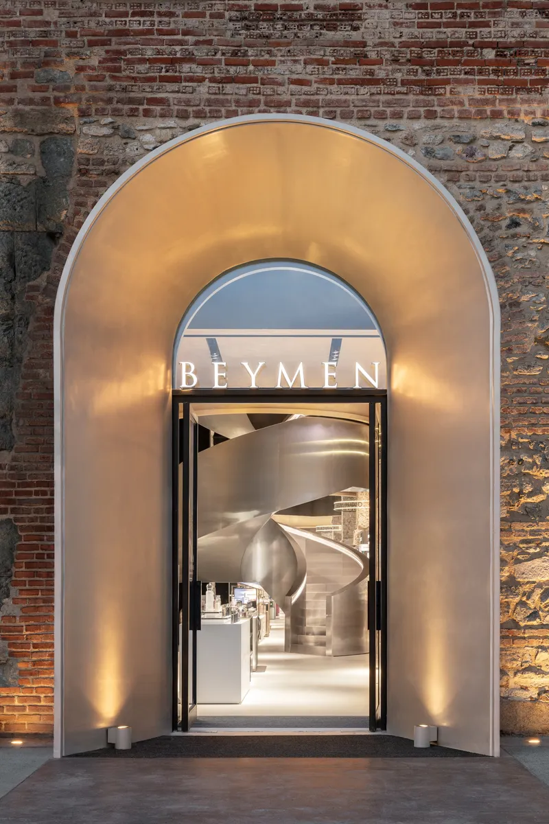 Backlit arched entry portal clad in polished metal against weathered brick and stone wall at dusk