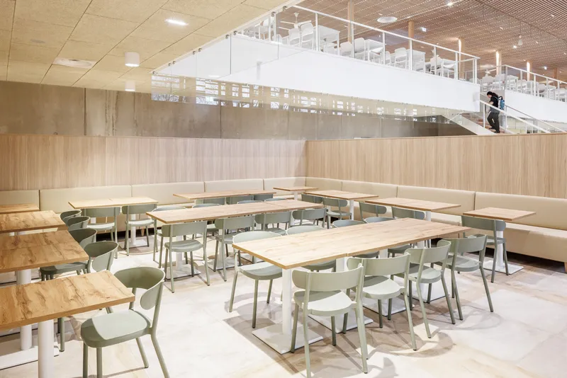 Dining area with banquette seating and light wood tables beneath a suspended white volume and slatted ceiling