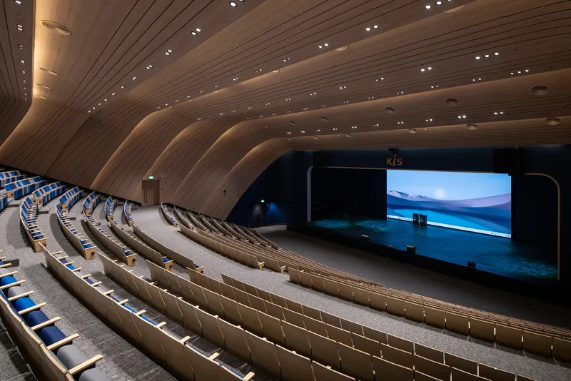 Auditorium interior with curved timber-clad ceiling and tiered seating facing an illuminated stage screen