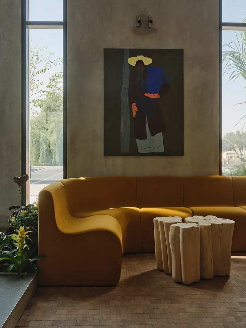 Yellow curved sofa with timber side tables beneath concrete walls and tall windows with greenery outside