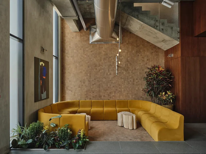 Double-height lobby with yellow sectional sofa beneath exposed concrete ceiling and ventilation ducts