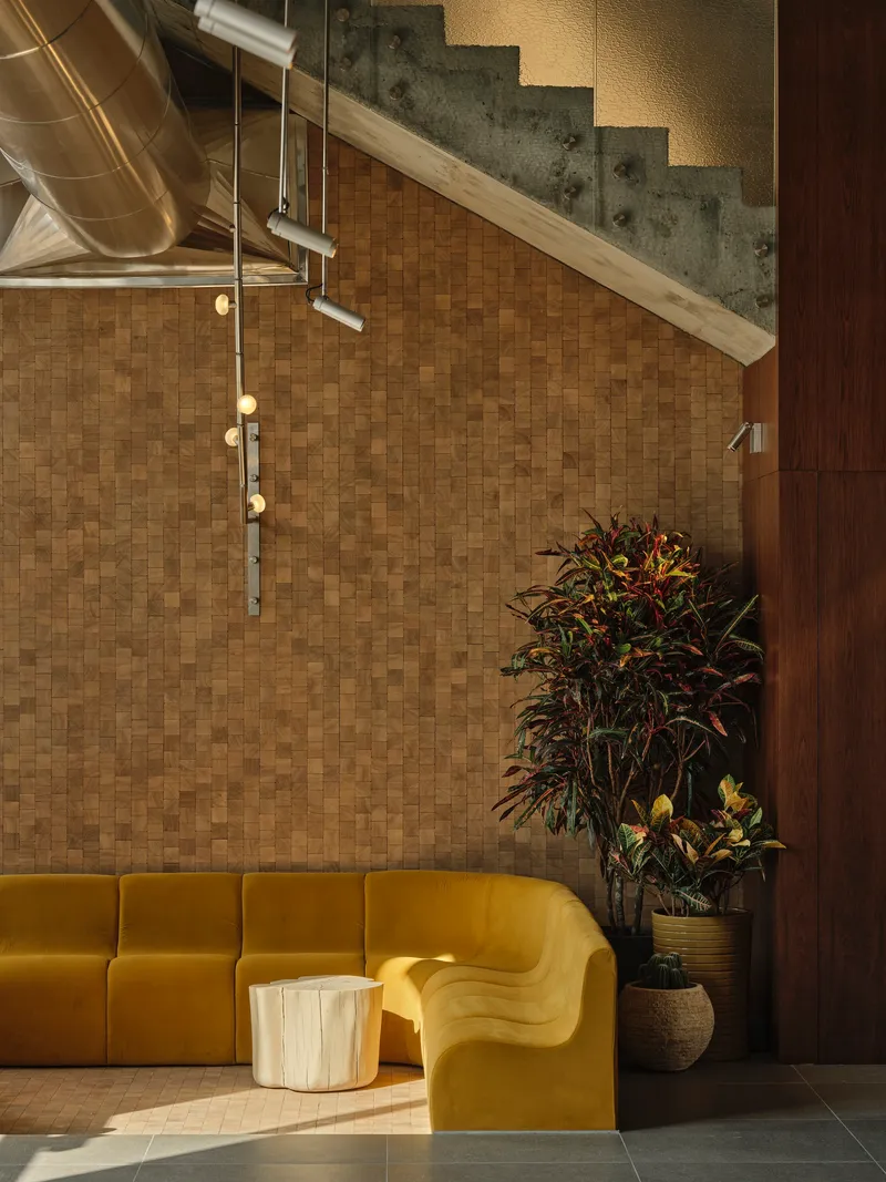Seating area with tiled cork wall under a concrete staircase lit by pendant fixtures and ductwork
