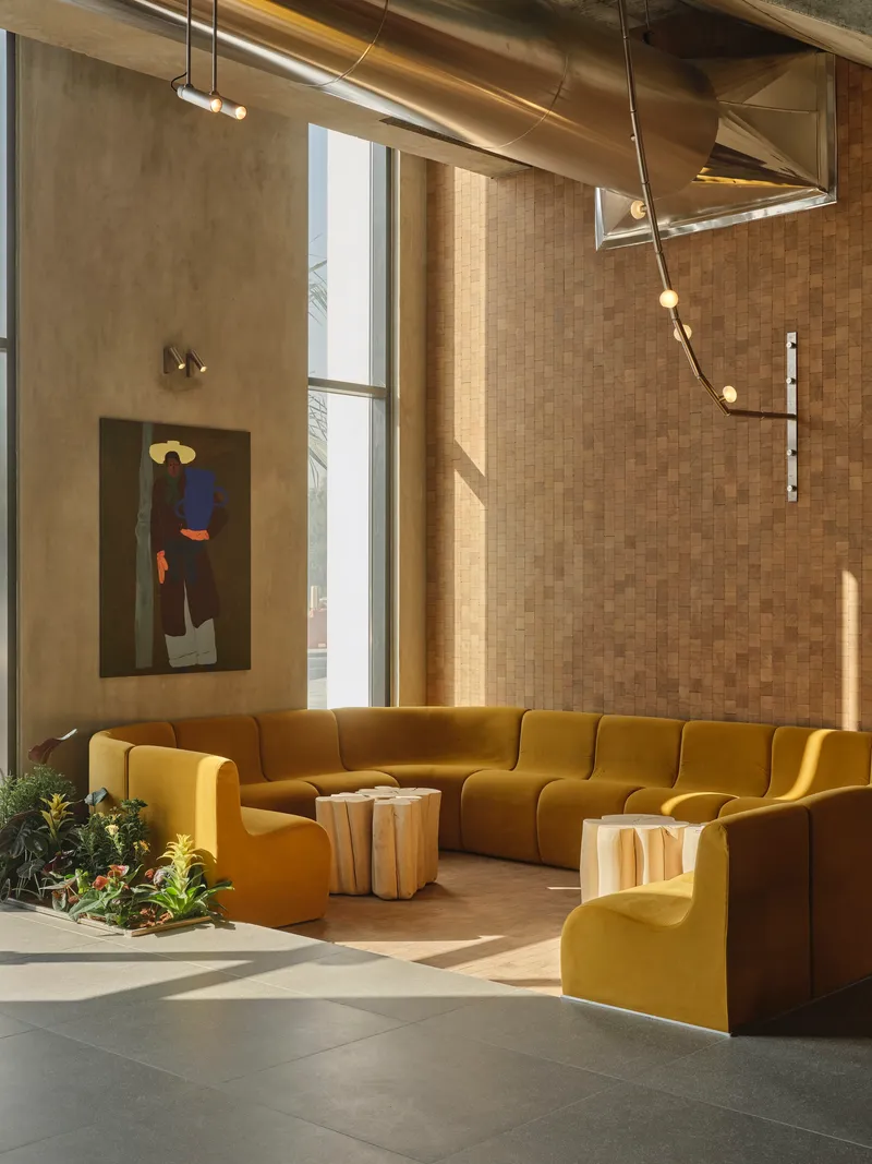 Double-height lounge with tall windows casting afternoon light across mustard upholstered seating and concrete floor