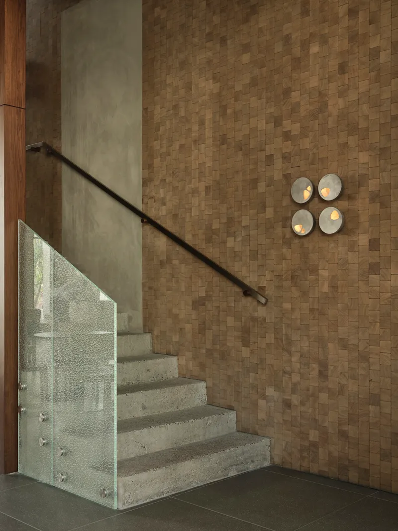 Concrete staircase with glass balustrade and metal handrail alongside a basketweave cork wall with circular sconces