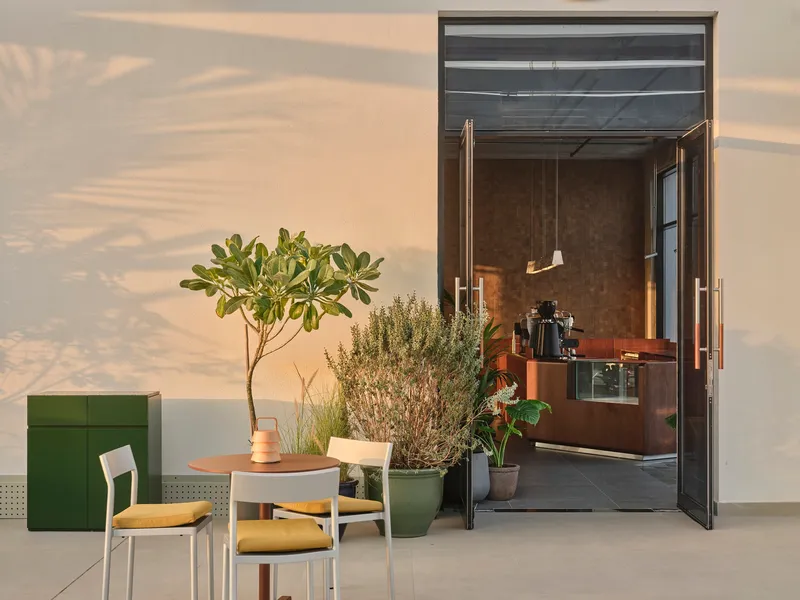 Outdoor seating area with potted plants and glass doors opening to a cafe interior in afternoon light
