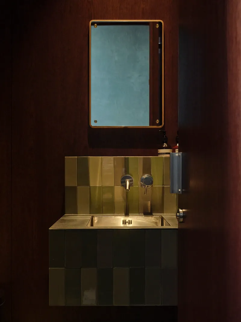Brass-framed mirror above a checkered tile backsplash and wall-mounted sink in a dark washroom