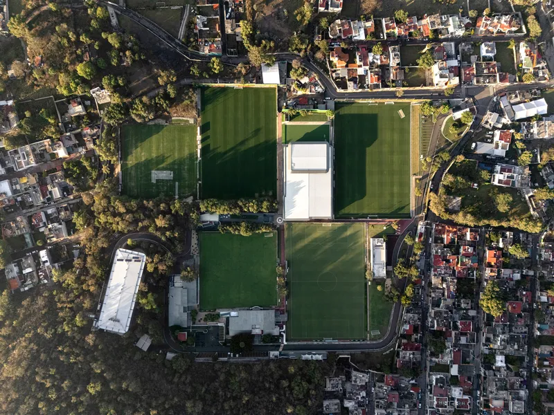Aerial view of athletic fields and white-roofed buildings surrounded by residential neighborhoods and wooded hillsides