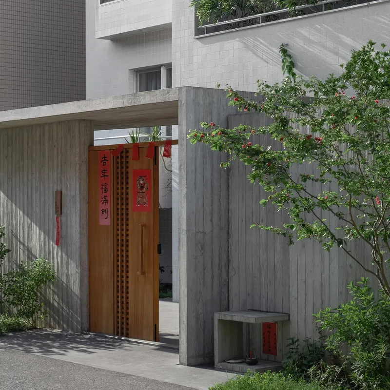 Entry courtyard with timber door and board-formed concrete walls beside a flowering tree casting shadows