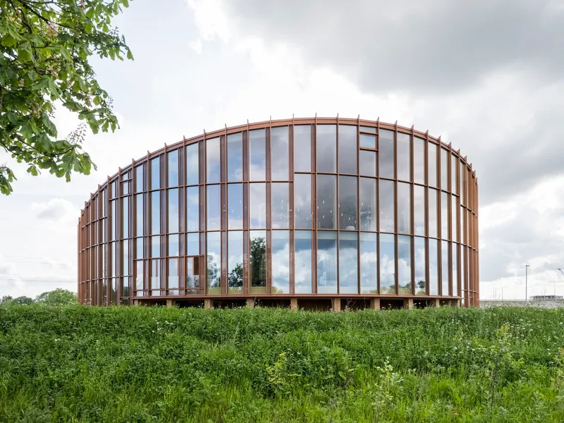 Cylindrical glass and timber facade rising from a meadow of tall grasses under cloudy sky
