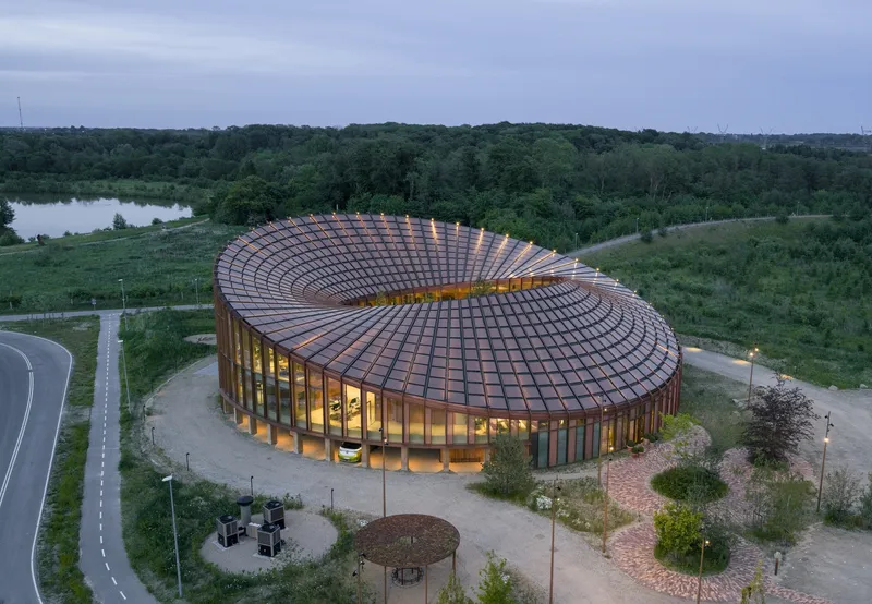 Aerial view of the circular building with spiraling copper tile roof at dusk next to forested landscape