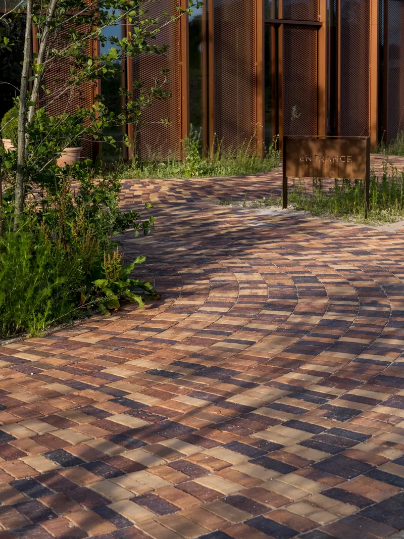 Multicolored brick paving path curving between planted beds with young saplings and bronze entrance sign