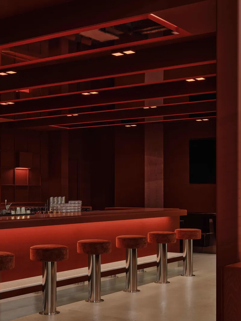 Bar counter with upholstered stools on metal pedestals beneath layered terracotta ceiling beams and recessed lighting