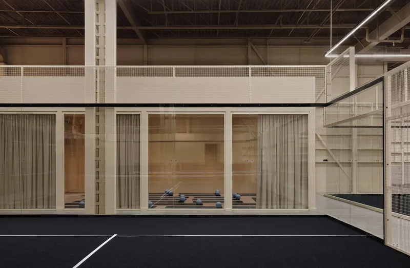 Interior sports court with glass-fronted viewing room, metal mesh partitions and exposed truss ceiling