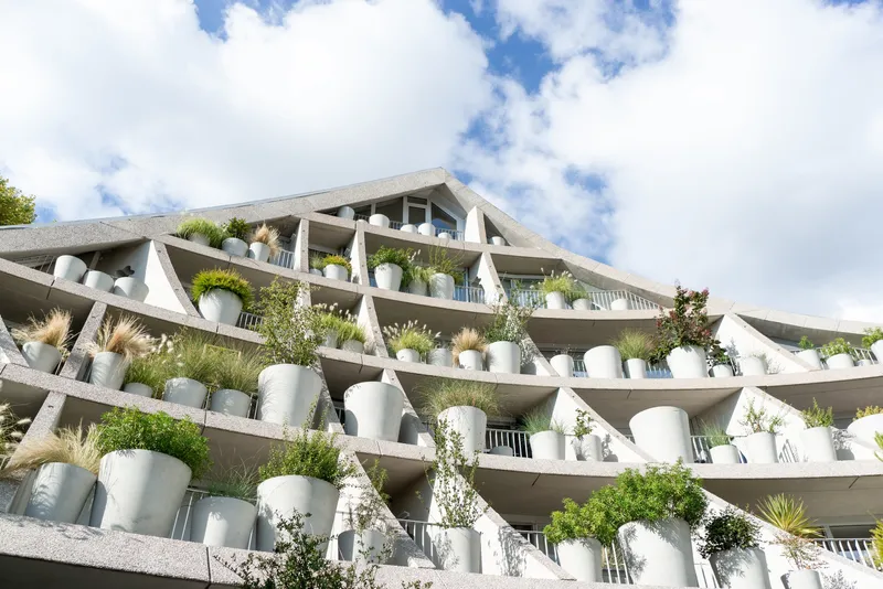 Stacked concrete terraces with white cylindrical planters filled with grasses and shrubs beneath a cloudy sky