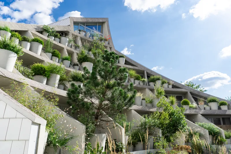 Tiered concrete facade with white cylindrical planters and vegetation cascading from each level under scattered clouds