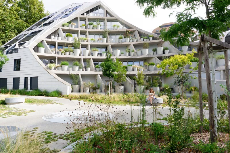Glazed gable end and planted balconies viewed across a landscaped plaza with grasses and timber arbor
