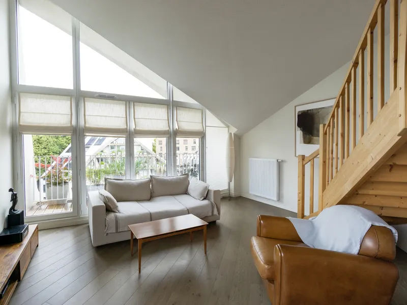 Living room with sloped ceiling, full-height windows, timber staircase and afternoon sunlight streaming in