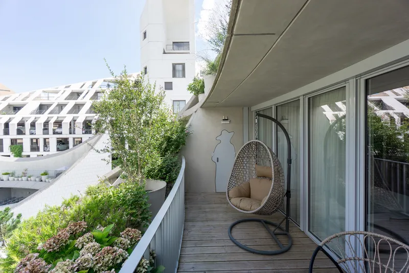 Timber deck balcony with hanging chair, planter bed, and concrete soffit overhead in daylight