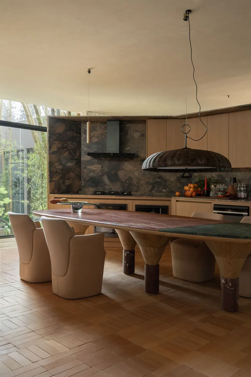 Sculptural dining table with curved timber legs below a woven dome pendant near glazed garden views