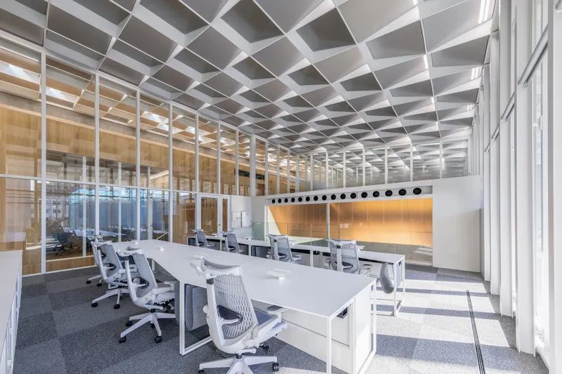 Open office workspace with white desks and chairs under a coffered ceiling grid with glass partitions