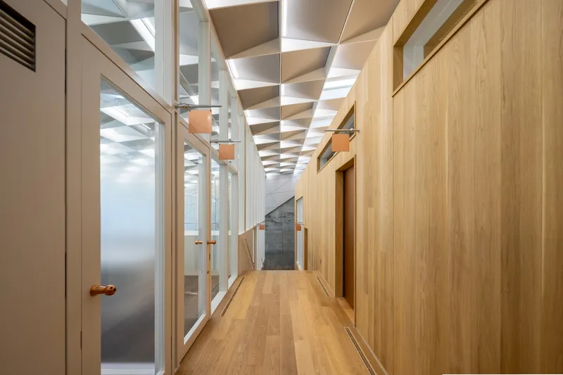 Wood floored corridor with timber wall paneling and white frame partitions under a geometric coffered ceiling