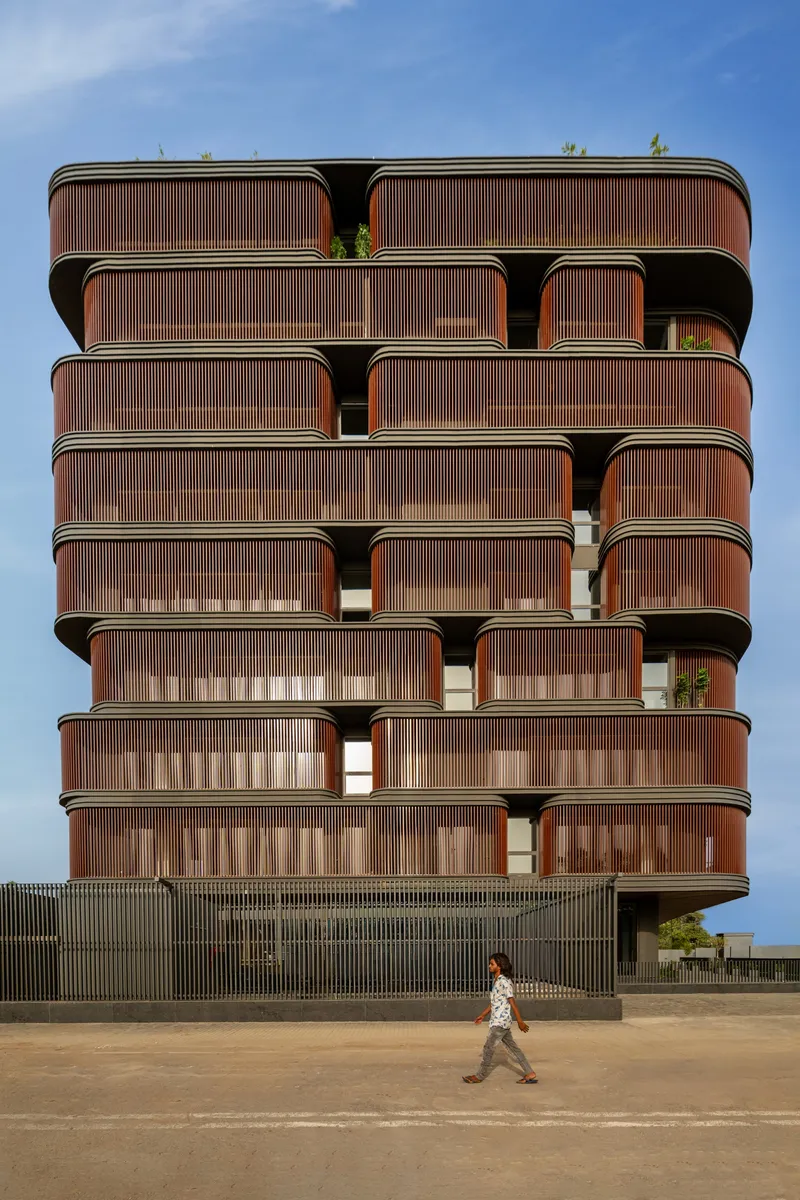 Stacked facade with vertical timber-slatted balconies and a person crossing the sandy forecourt below