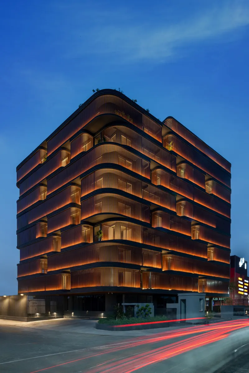 Corner view of the illuminated facade with curved balconies and bronze louvers at dusk with traffic trails