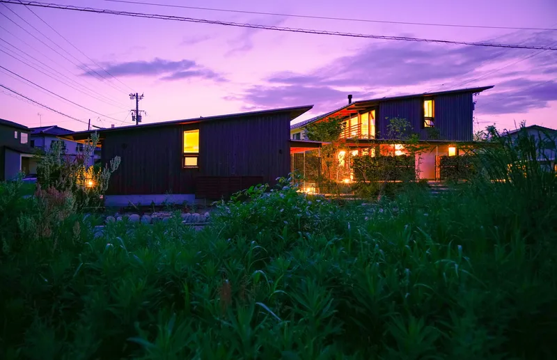 Dark timber-clad residence with illuminated windows viewed across a field of tall grasses at twilight