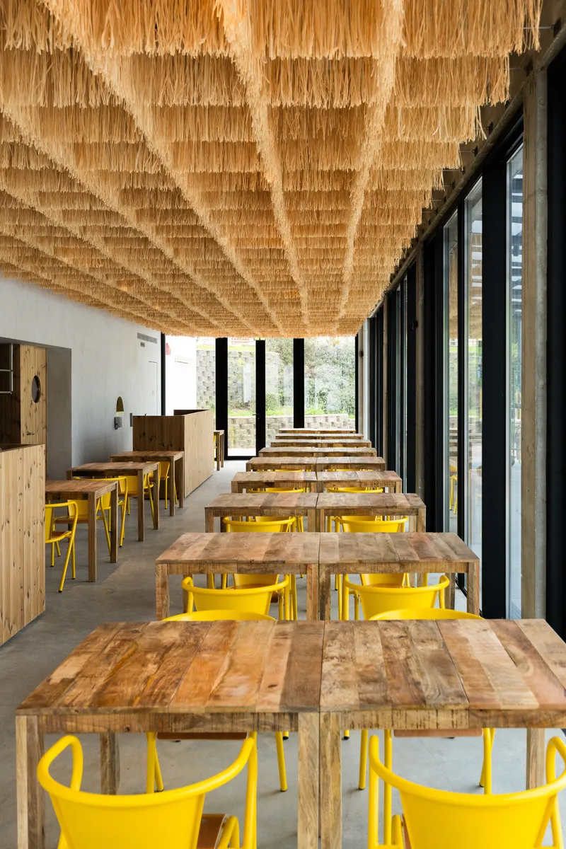 Long view through the dining room with repeated timber tables and yellow chairs under raffia canopy