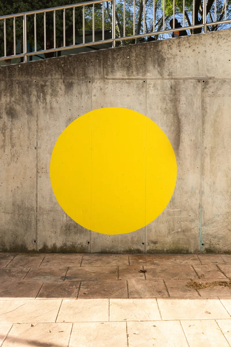 Yellow circle painted on a weathered concrete retaining wall below a steel handrail and trees
