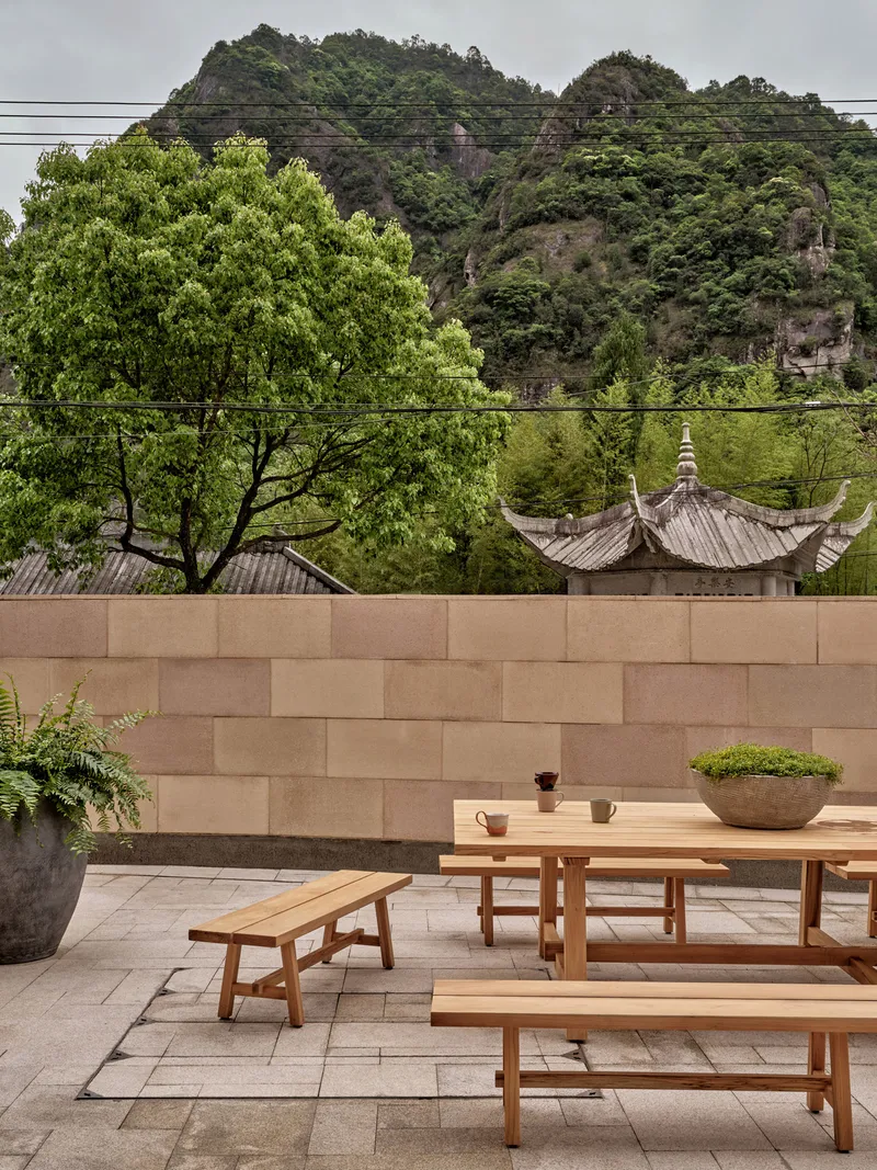Timber dining table and benches on paved terrace against sandstone wall with hillside beyond