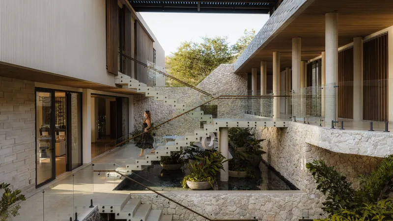 Glass staircase spanning courtyard pond between stone-clad volumes with timber ceiling overhead