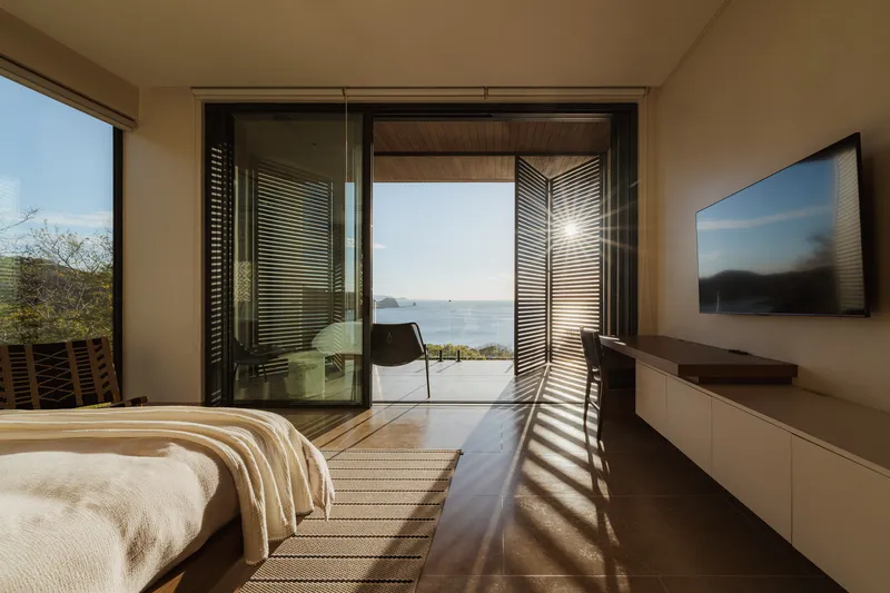 Bedroom with open sliding doors framing an ocean view and casting louvered sunlight patterns