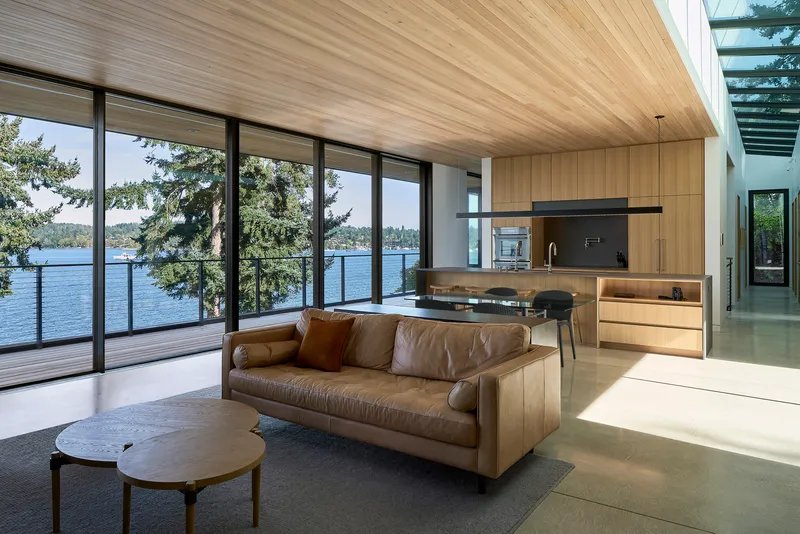 Open living space with timber ceiling and floor-to-ceiling glazing overlooking a lake through evergreen trees