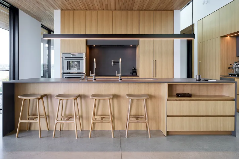 Kitchen island with four timber stools beneath a suspended wood-clad volume and linear pendant light