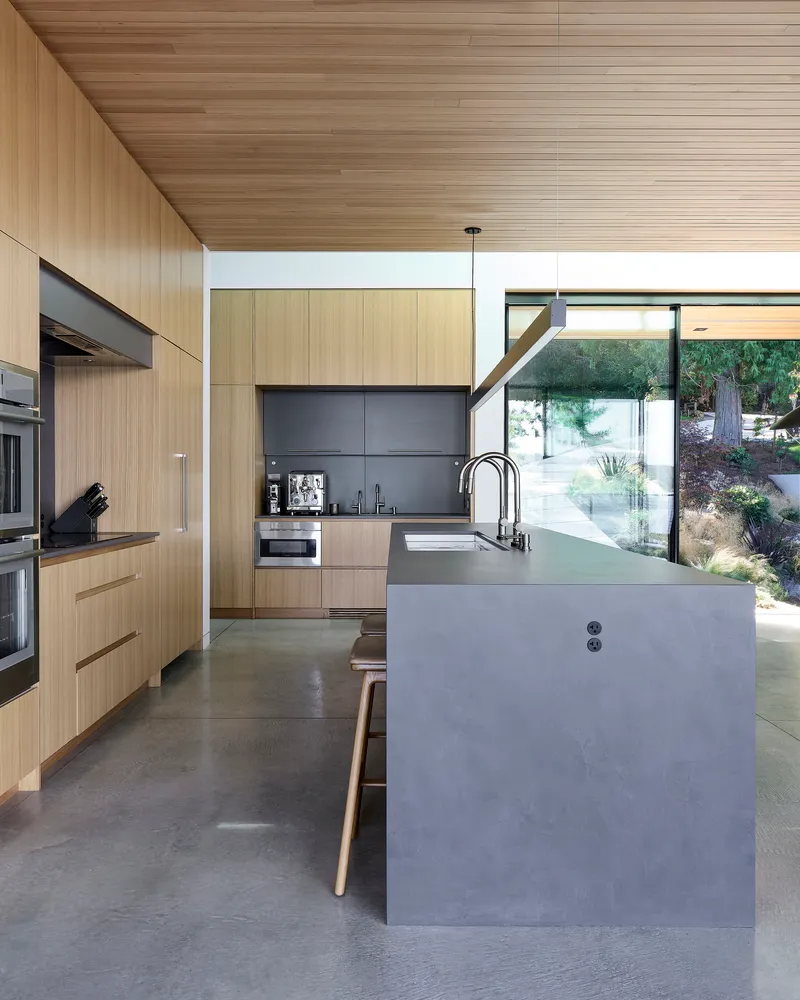 Kitchen island with pendant light fixture beneath wood-paneled ceiling overlooking a sloped garden through glazed walls