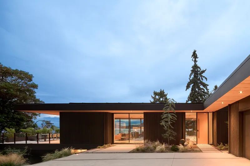 Courtyard entry with dark vertical cladding, timber soffit, and planted beds at dusk