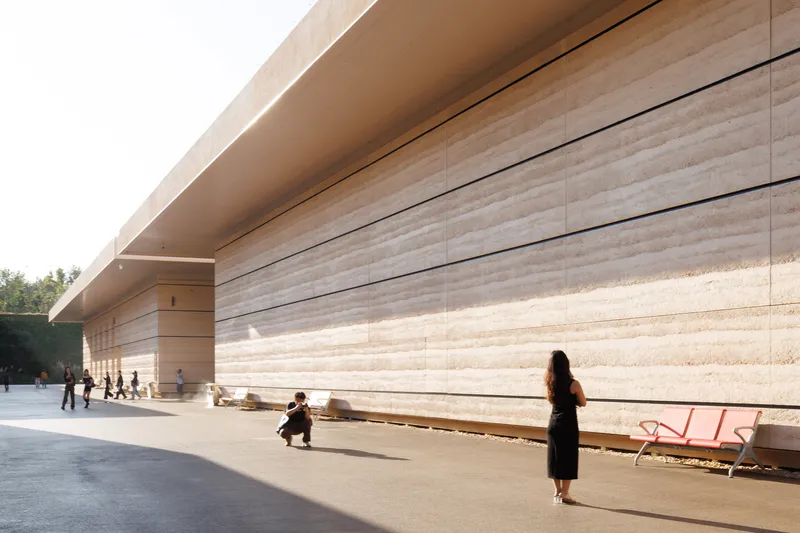 Long travertine facade with horizontal banding alongside a paved plaza with visitors and red benches in afternoon light