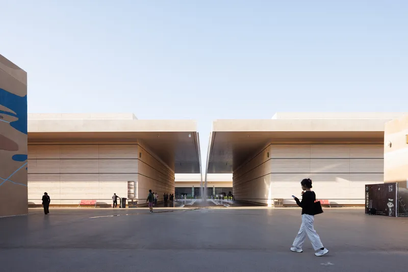 Courtyard plaza with two banded travertine volumes flanking a central slot under a pale sky