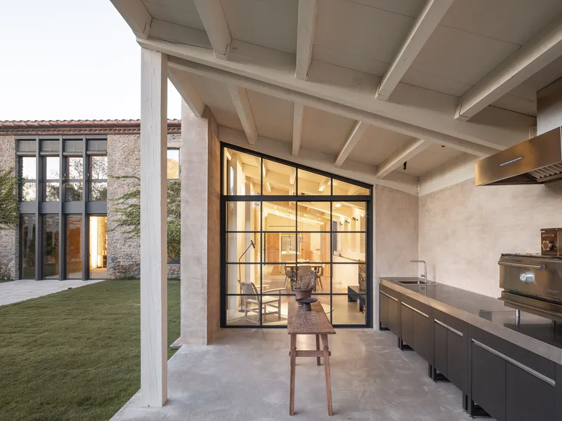 Covered outdoor kitchen with exposed beam ceiling and black-framed glass doors opening to an interior dining area