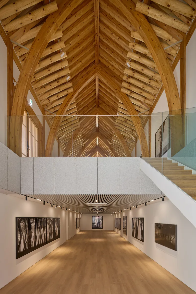 Gallery space below a vaulted timber ceiling with concrete walls and framed photographs under track lighting