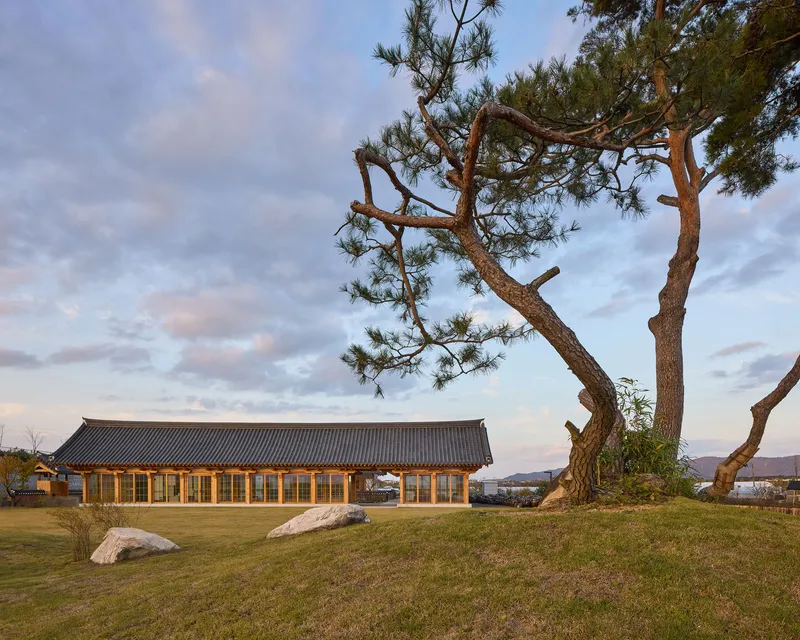 Long timber pavilion with tiled roof beneath pine trees on a grassy slope at dusk