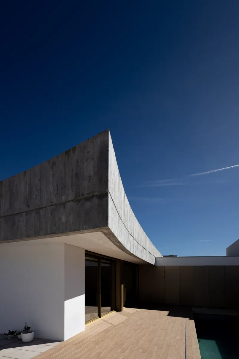 Timber deck terrace adjacent to a swimming pool beneath the angular concrete roof edge
