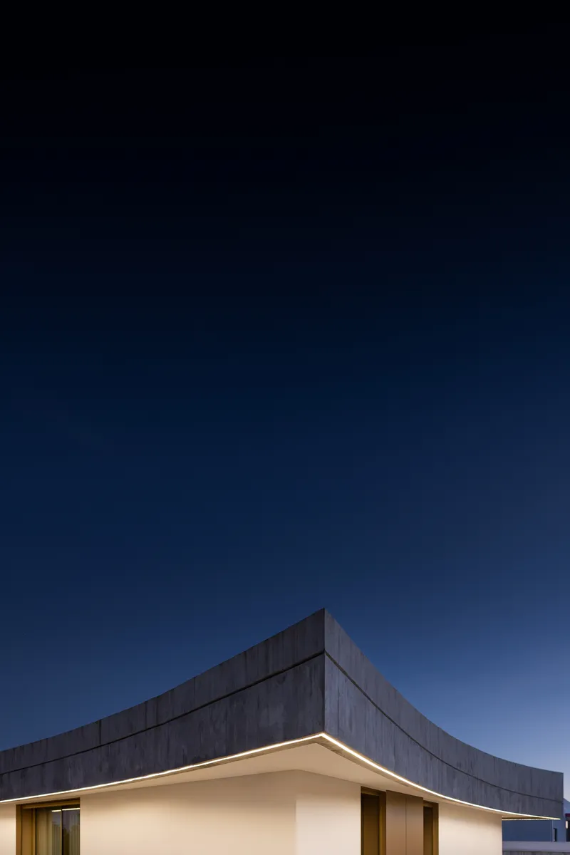 Angular concrete upper volume rising above the illuminated white plaster base at dusk