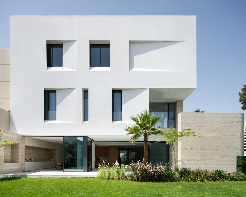 Garden facade showing cantilevered white volumes above travertine base and planted lawn with palm
