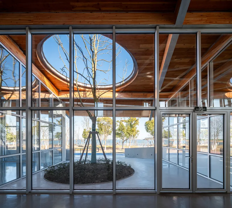 Interior view through glazed curtain wall toward circular planted courtyard with bare trees under timber ceiling