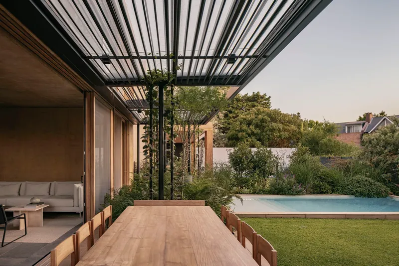 Outdoor dining table beneath slatted steel canopy with pool and lush planting beyond